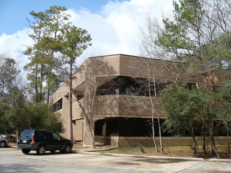 A two-storey brown brick office building with bands of bronze reflective windows, seen from its car park on a clear day. Pines and bare hardwood trees stand around it, and marked parking bays run along the front with a car parked in one.